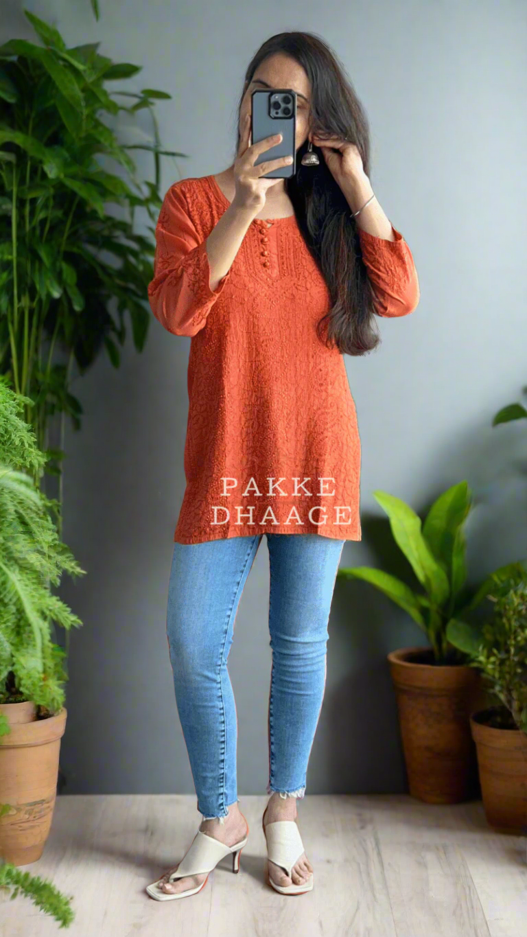 Person taking a mirror selfie wearing an orange chikankari top and blue jeans in a room with plants.