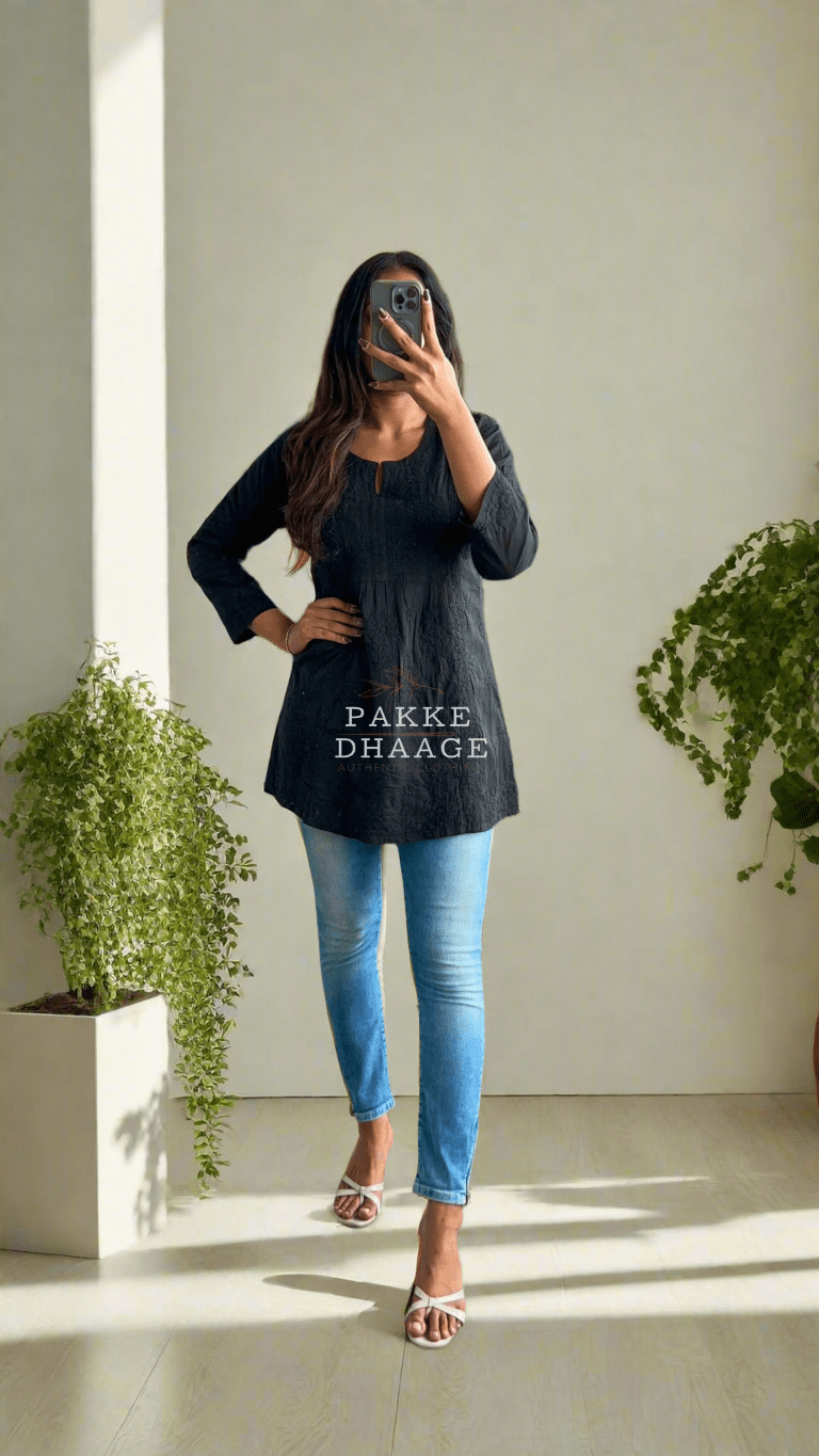 A person wearing a classic black short kurti with Chikankari embroidery, paired with blue jeans, standing in a room with a lot of plants.