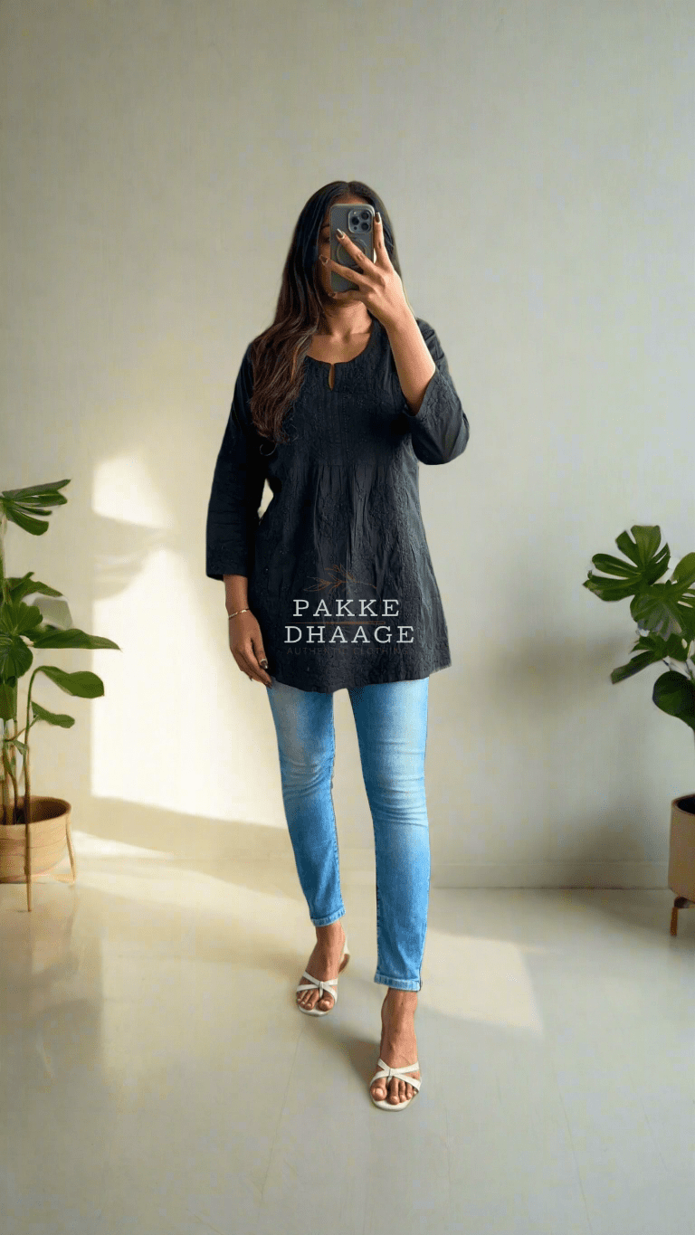 Person taking a mirror selfie wearing a black top and blue jeans in a room with plants.