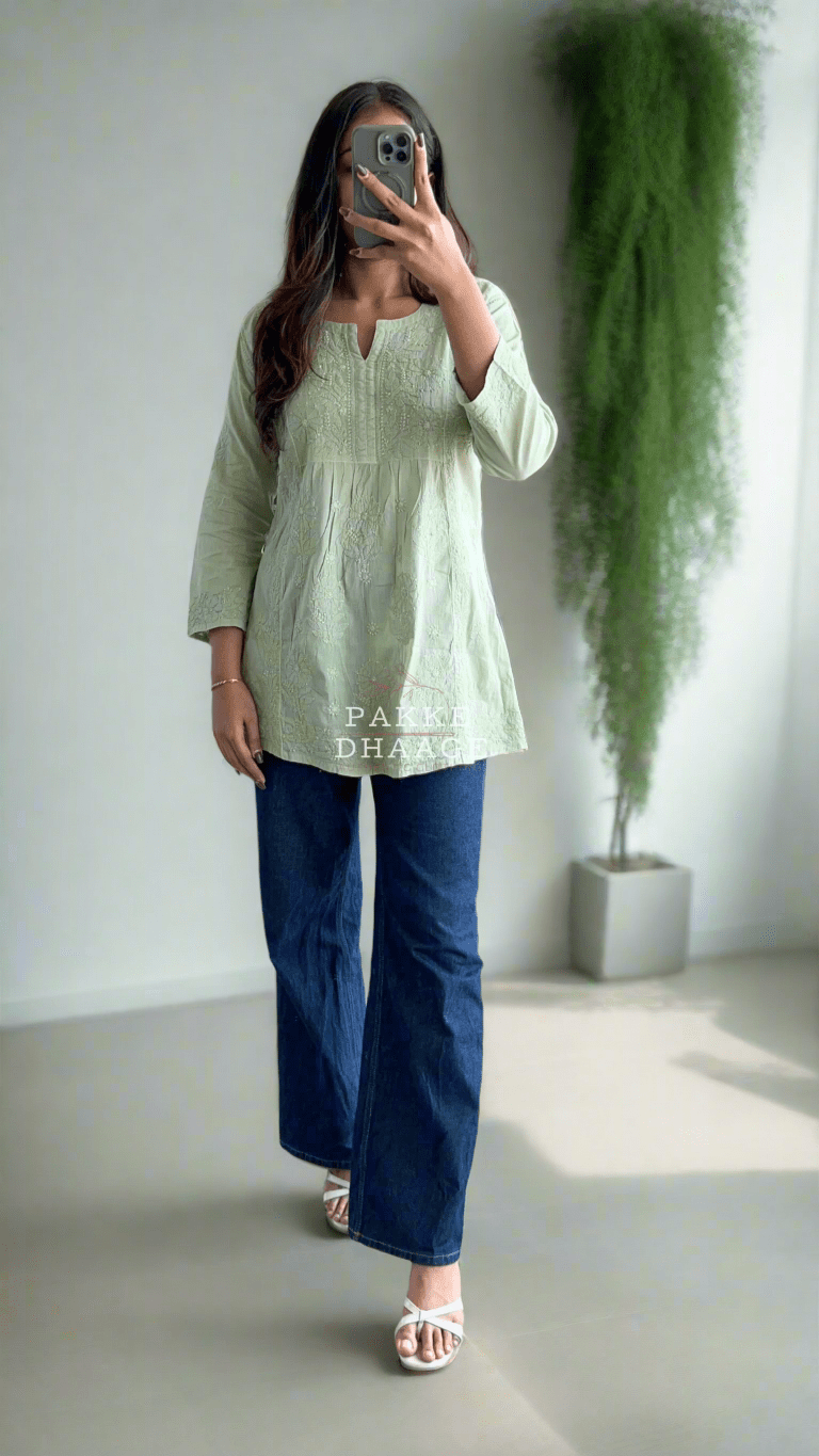 Person taking a mirror selfie wearing a light green top and blue pants on a balcony with plants.