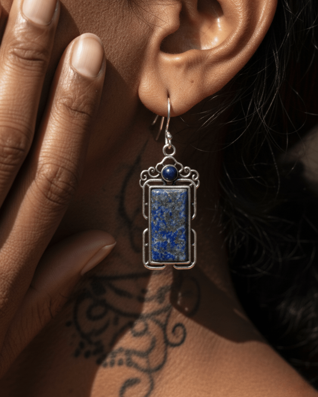 Earring with blue stone and silver frame worn by a person with tattoos on their arm.