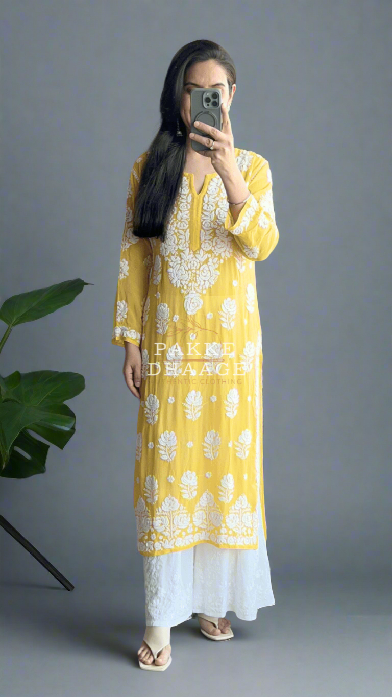 Person wearing a yellow and white chikankari kurti taking a mirror selfie in a plant-filled room.