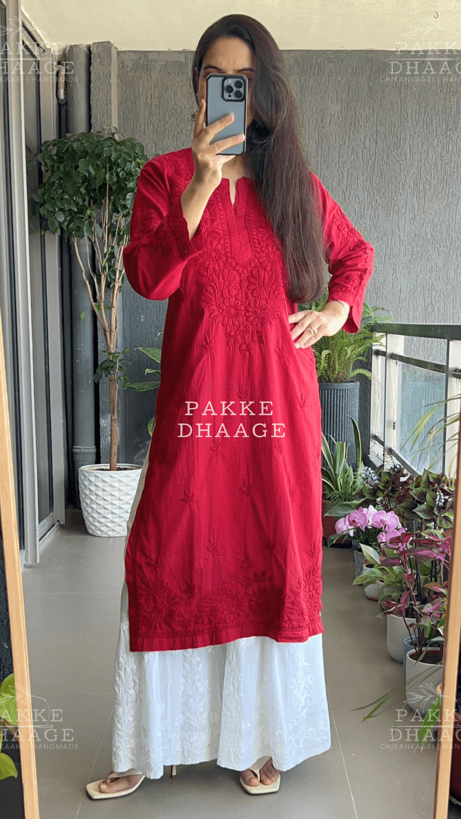 Person wearing a red chikankari kurta standing in front of a mirror with plants in the background.