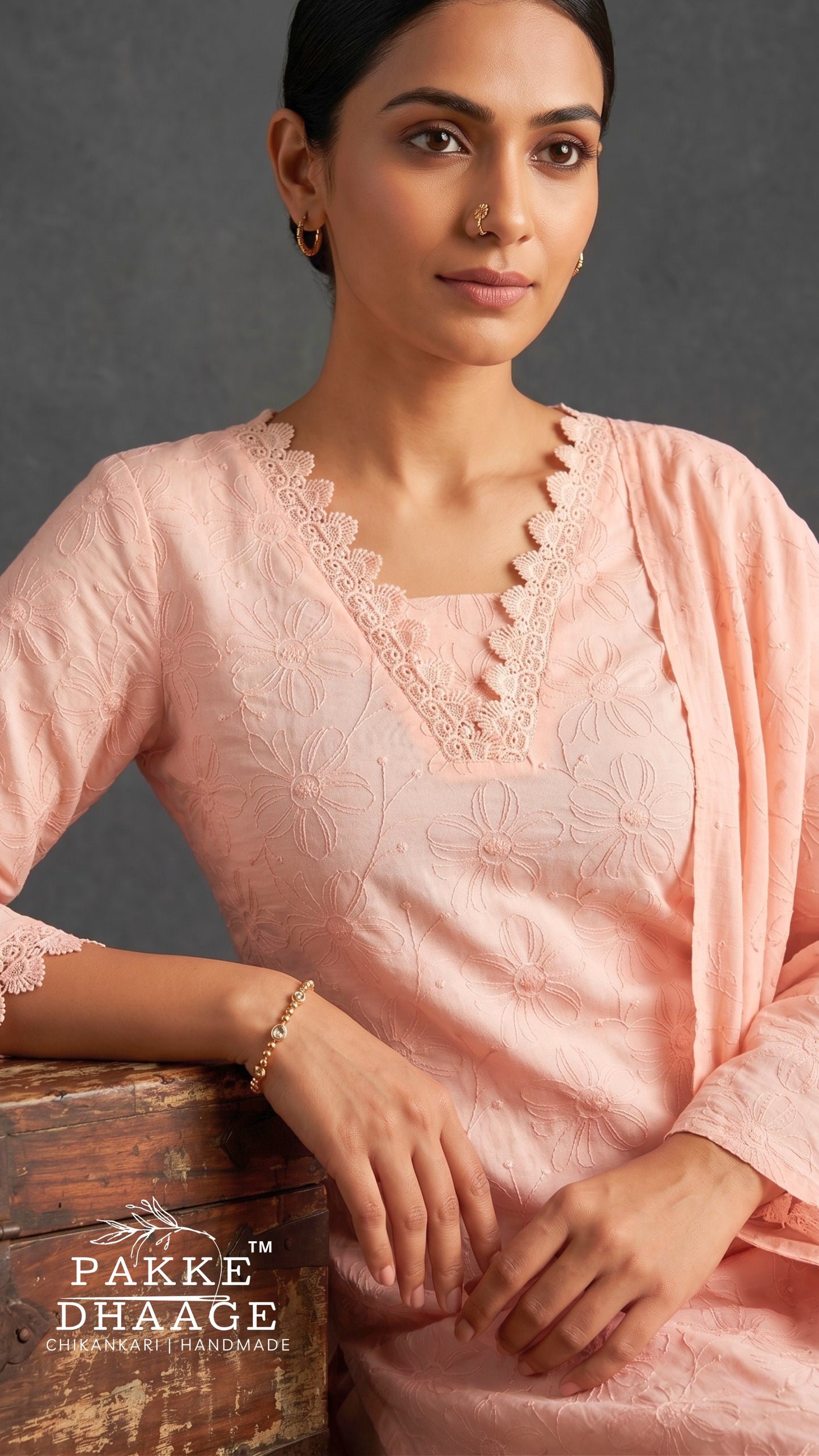 A woman wearing a light pink embroidered kurta with scalloped lace trim at the V-neck, paired with a matching draped scarf. The brand logo 'PAKE DHAAGE' is visible in the bottom-left corner over a rustic wooden surface.