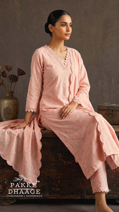 A woman wearing a light pink three-piece salwar kameez set sits on a rustic wooden chest against a textured gray wall. The set includes a long-sleeve tunic with scalloped edges, matching trousers, and a draped dupatta.