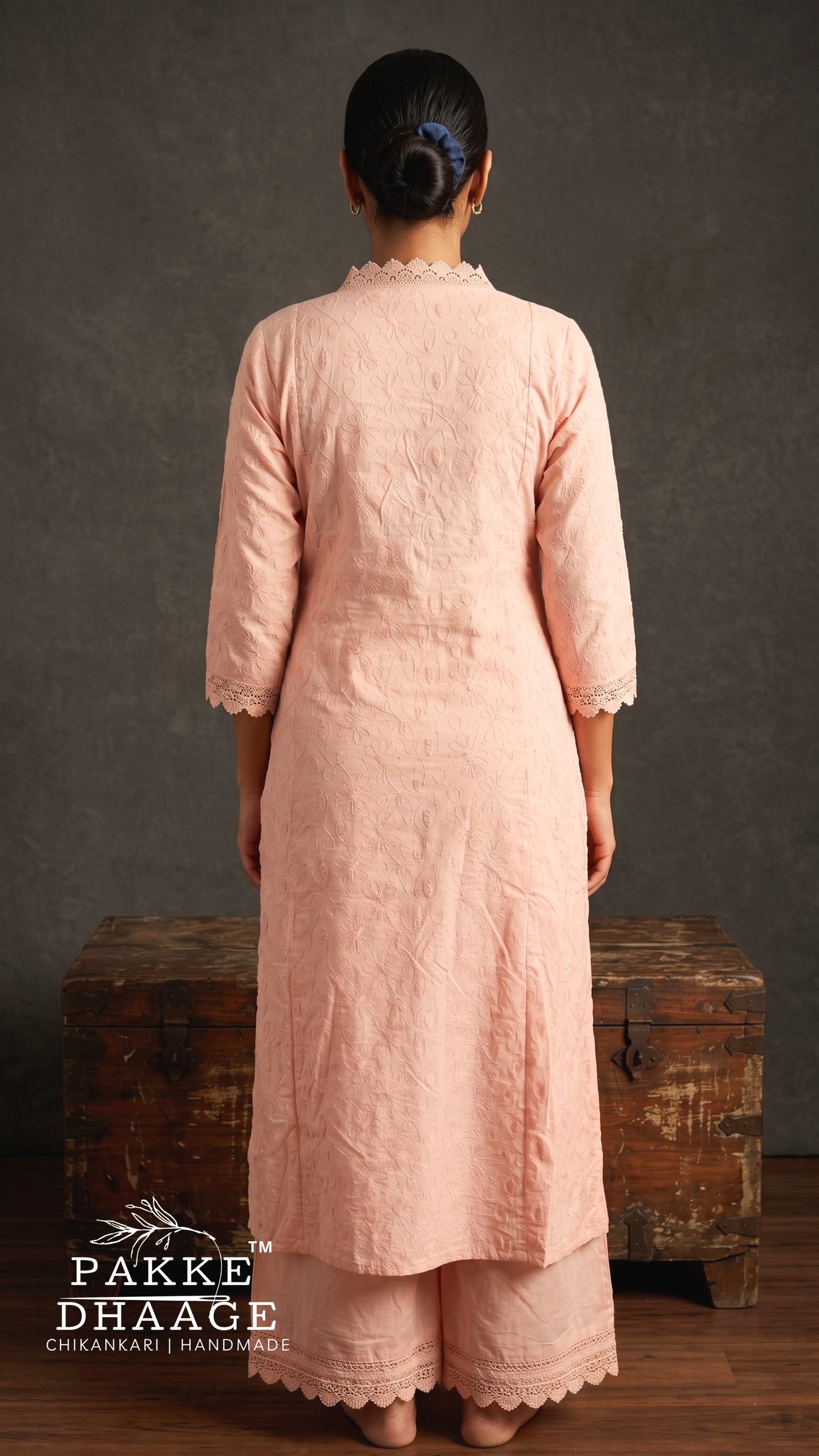 A woman viewed from behind wearing a light peach-pink three-piece chikanari kurta and trouser set with scalloped edges, standing in front of a rustic wooden chest against a textured gray wall.
