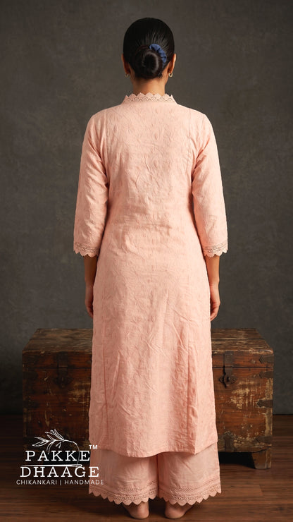 A woman viewed from behind wearing a light peach-pink three-piece chikanari kurta and trouser set with scalloped edges, standing in front of a rustic wooden chest against a textured gray wall.