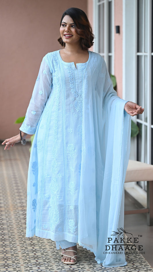 A woman modeling a light blue Chikankari embroidered kurta set with a matching dupatta, standing on a patterned tile floor in front of a building entrance. The brand name 'PAKKE DHAAGE' is visible as a watermark in the bottom right corner.