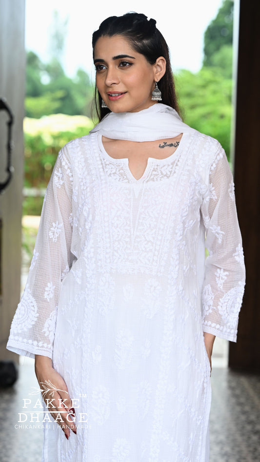 A woman wearing a white chikankari embroidered kurta with sheer sleeves and a matching white dupatta, photographed in a softly lit indoor setting with blurred greenery in the background. A watermark reading 'PAKKE DHAAGE' appears at the bottom left.