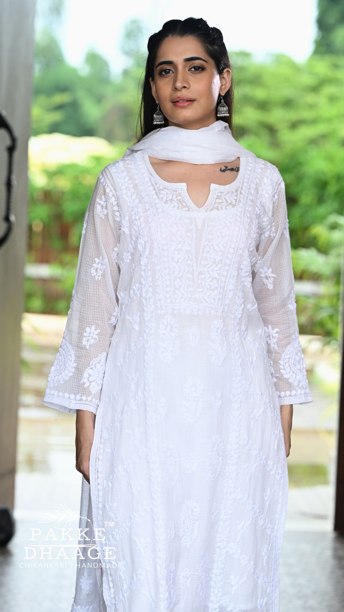 A woman wearing a white chikankari kurta with sheer sleeves and a matching white dupatta, standing in a doorway with a blurred green outdoor background. A 'Pakke Dhaage' watermark is visible in the bottom left corner.