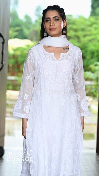 A woman wearing a white chikankari kurta with sheer sleeves and a matching white dupatta, standing in a doorway with a blurred green outdoor background. A 'Pakke Dhaage' watermark is visible in the bottom left corner.