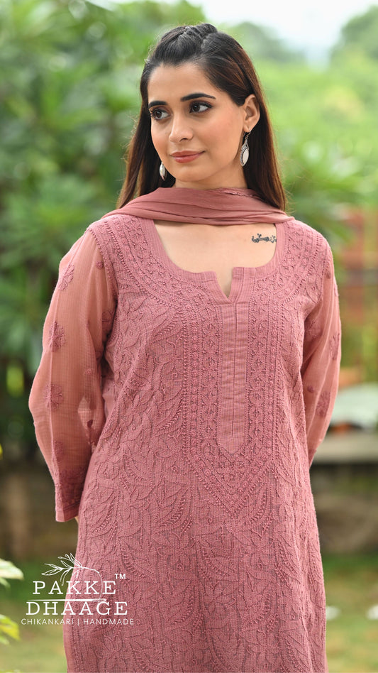 A woman wearing a chocolate chikankari embroidered kurta with a matching dupatta, standing outdoors against a blurred green garden background. The brand logo 'Pakke Dhaage' is visible in the bottom left corner.