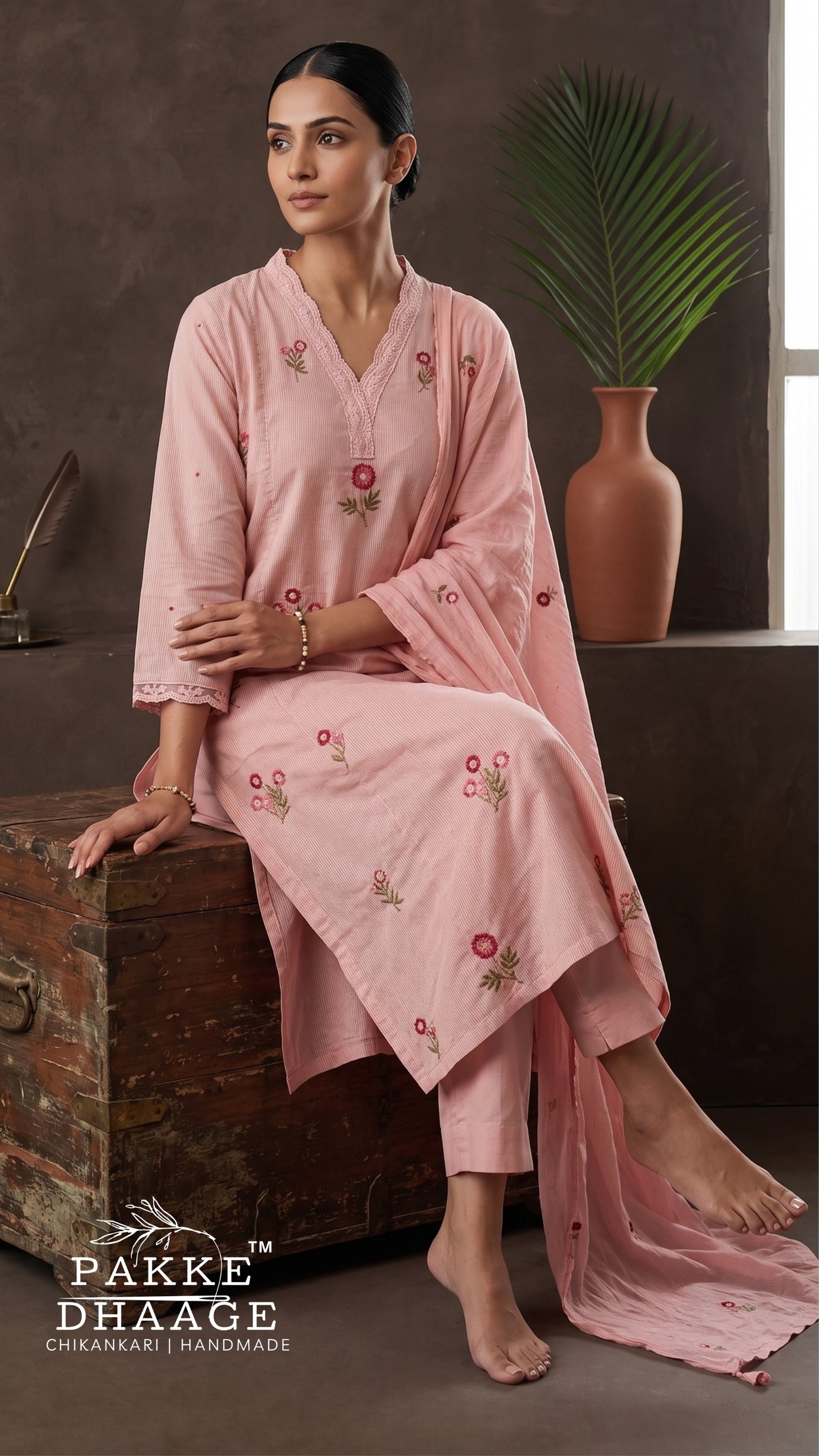 A woman models a light pink ethnic kurta set with floral embroidery, sitting on a rustic wooden chest in a styled interior setting. The set includes a long-sleeved kurta, matching trousers, and a dupatta.