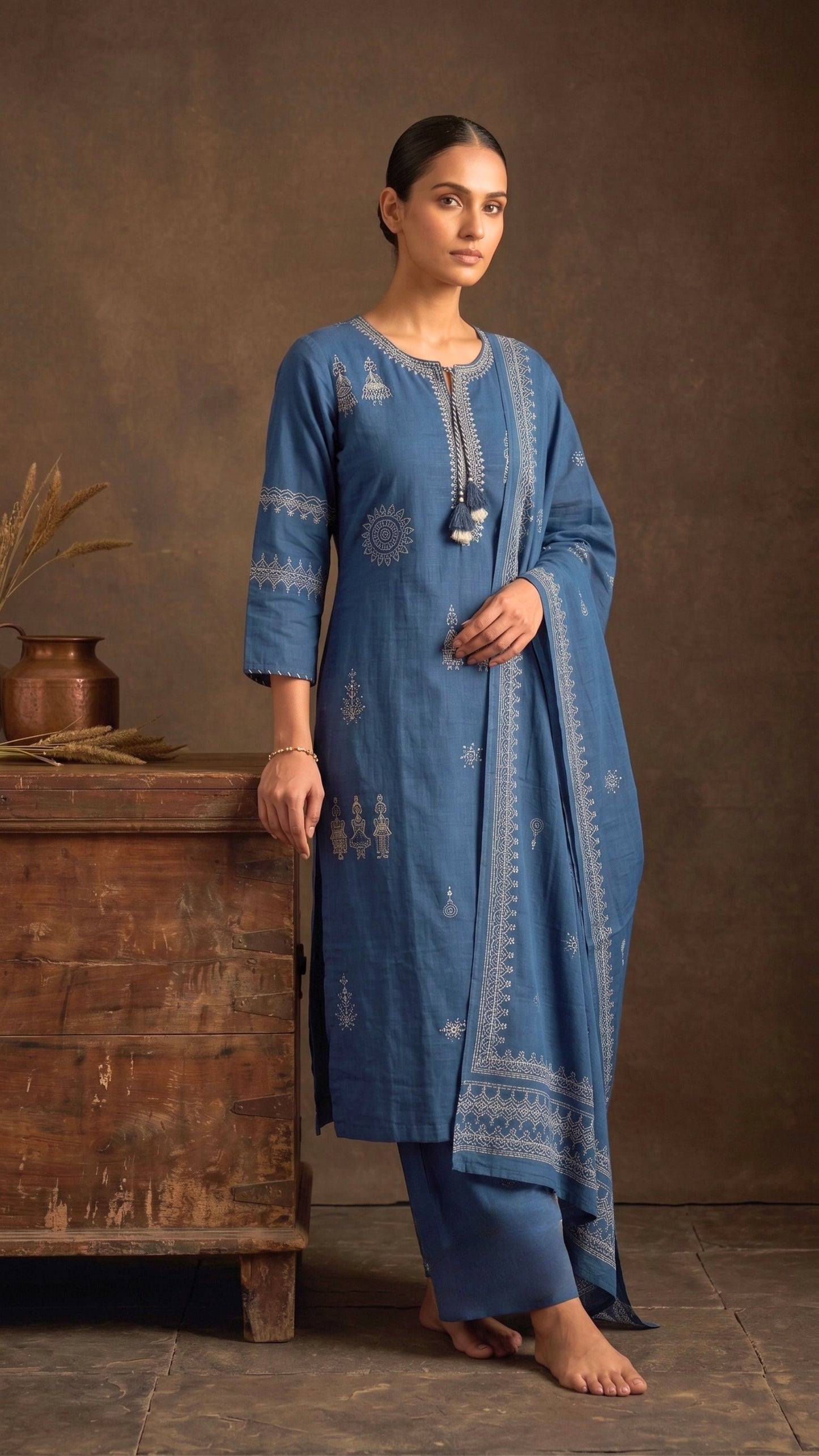 A woman models a blue three-piece salwar kameez suit with white printed motifs, standing barefoot next to a rustic wooden chest against a textured brown backdrop. Pakke Dhaage.
