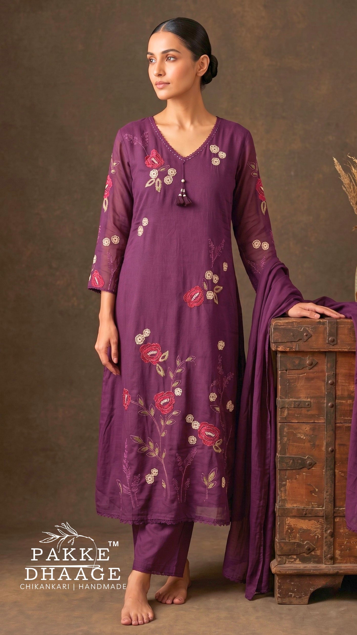 Model standing leaning on a trunk wearing a deep plum purple pure cotton straight kurta set with vibrant floral vine embroidery, scalloped lace V-neck with tassel, semi-sheer sleeves, matching pants, and dupatta.