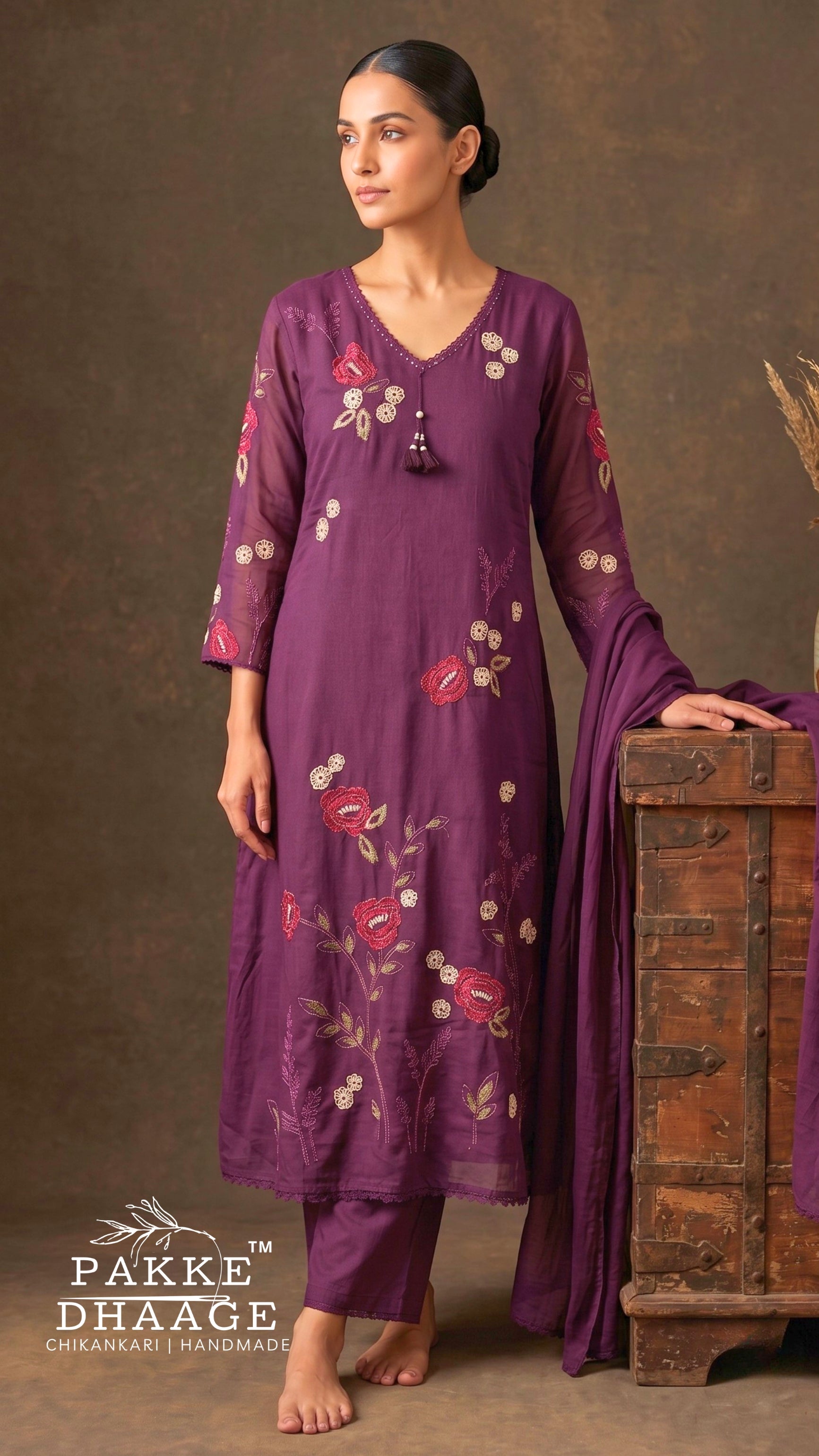 Model standing leaning on a trunk wearing a deep plum purple pure cotton straight kurta set with vibrant floral vine embroidery, scalloped lace V-neck with tassel, semi-sheer sleeves, matching pants, and dupatta.