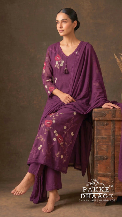 A woman models a purple three-piece salwar kameez suit with floral embroidery, sitting on a rustic wooden chest against a textured brown backdrop. The brand logo 'Pakke Dhaage' is visible in the bottom right corner.