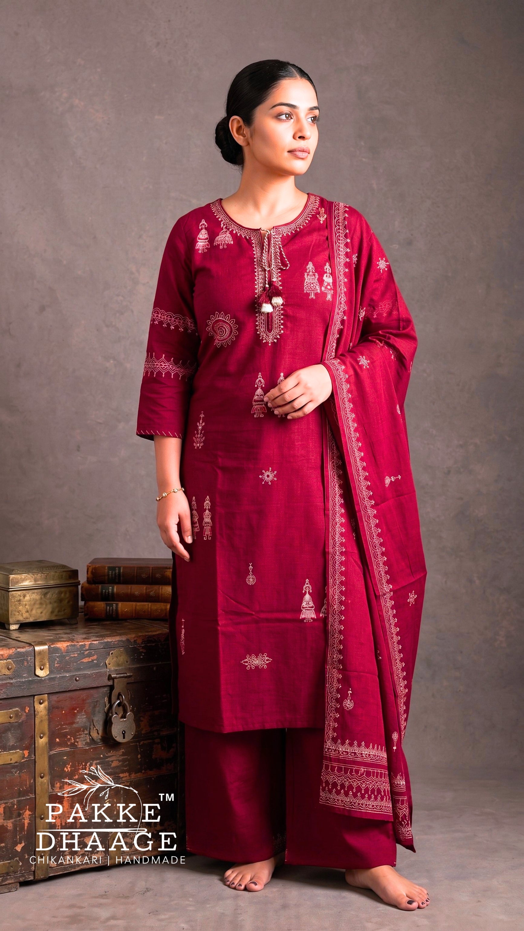 A woman modeling a deep maroon three-piece salwar kameez suit with matching dupatta, featuring intricate gold and white embroidery, set against a textured gray studio background with a vintage wooden chest. | PAKKE DHAAGE