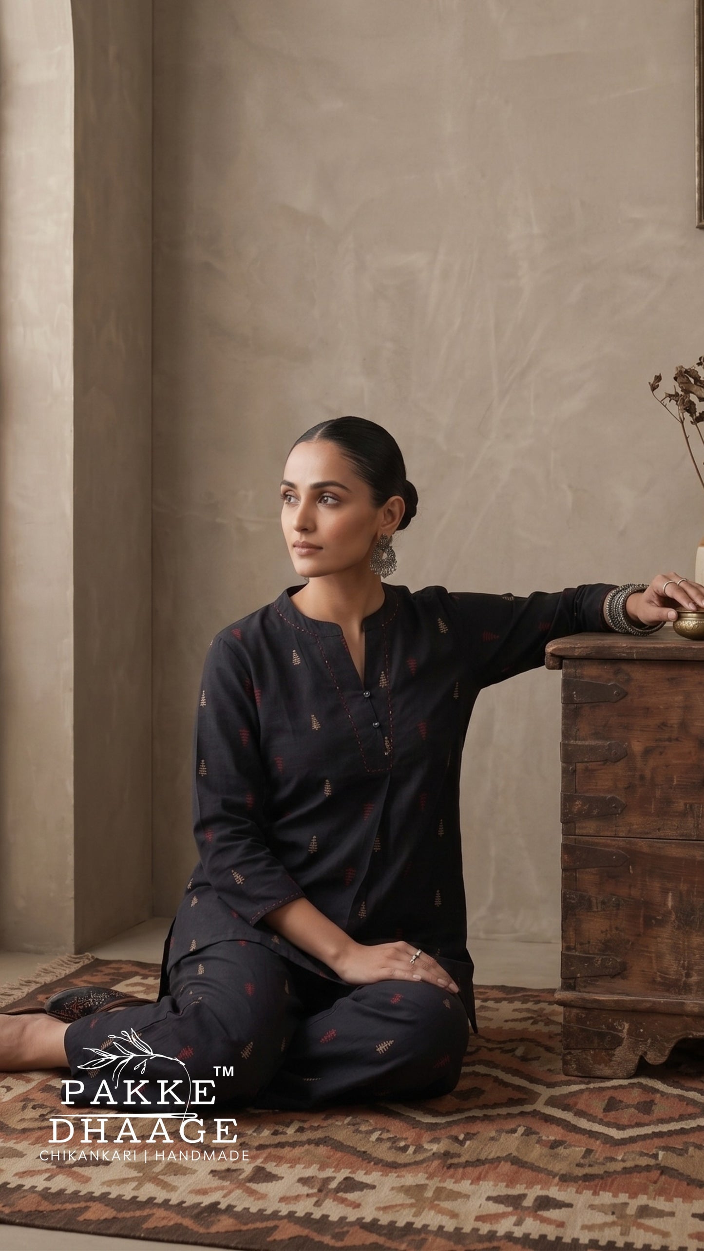 A woman sits cross-legged on a patterned rug in a rustic indoor setting, wearing a charcoal black kurta and matching pants with subtle embroidered details. She is posed next to a wooden chest, with a textured beige wall behind her.