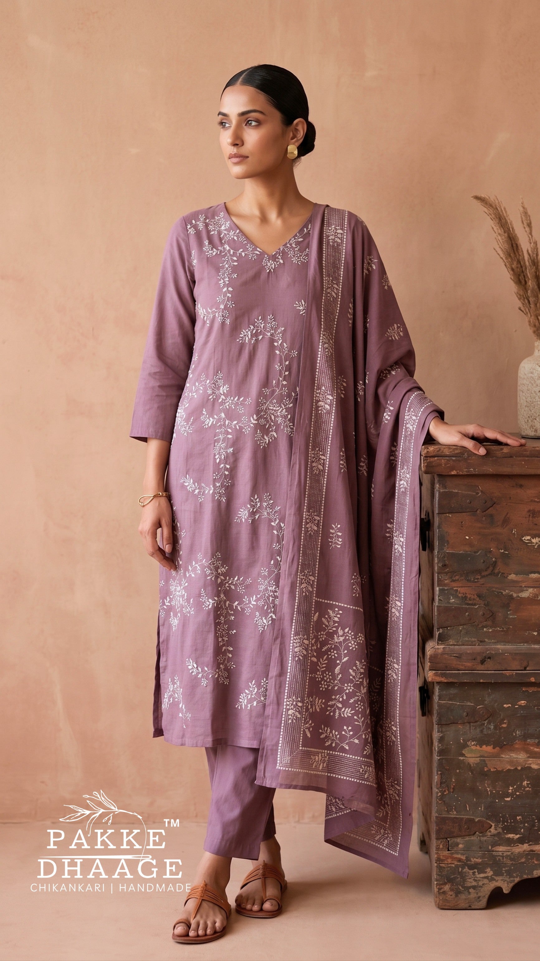 A woman models a three-piece dusty purple ethnic suit with white floral embroidery, consisting of a kurta, pants, and a dupatta, posed next to a rustic wooden chest against a warm beige background.