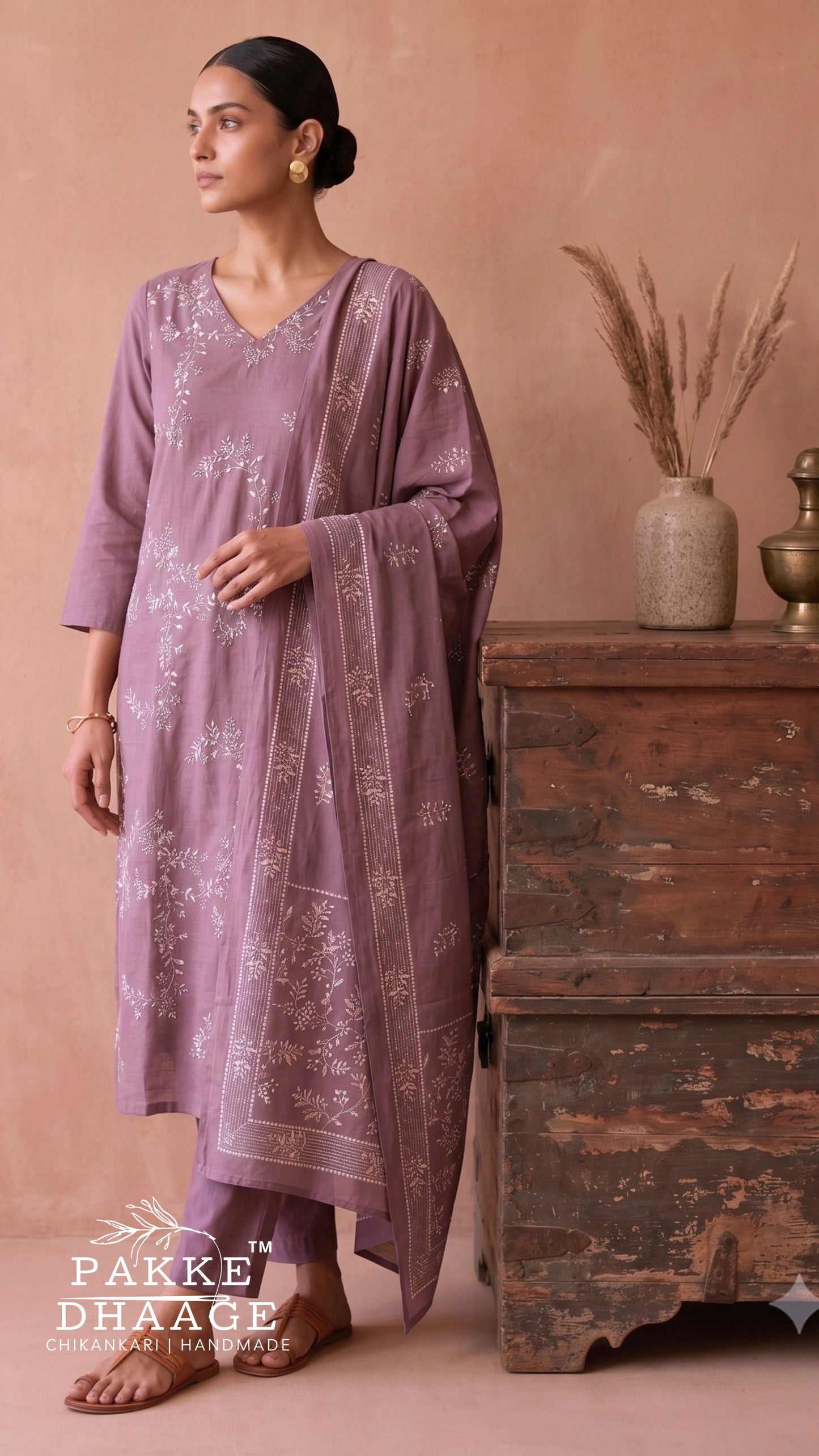 A woman models a lavender purple ethnic kurta set with matching dupatta and trousers, featuring delicate white floral embroidery. She is standing in a rustic room next to a weathered wooden chest, with a warm beige wall in the background.