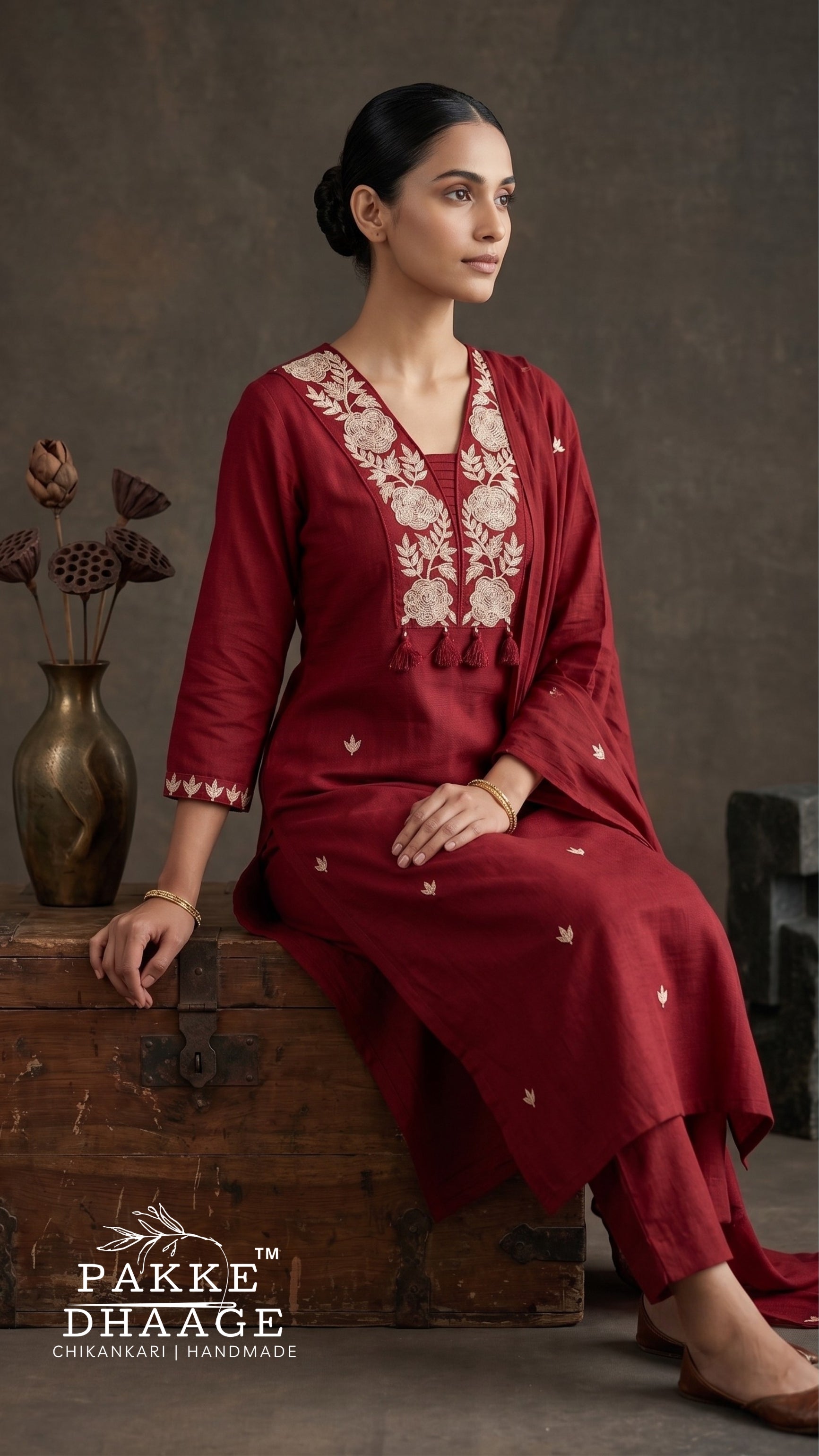 A woman models a deep red salwar kameez suit with beige floral embroidery on the neckline and sleeves, sitting on a rustic wooden crate against a textured brown studio backdrop. The brand logo 'PAKE DHAGE' is visible in the bottom-left corner.
