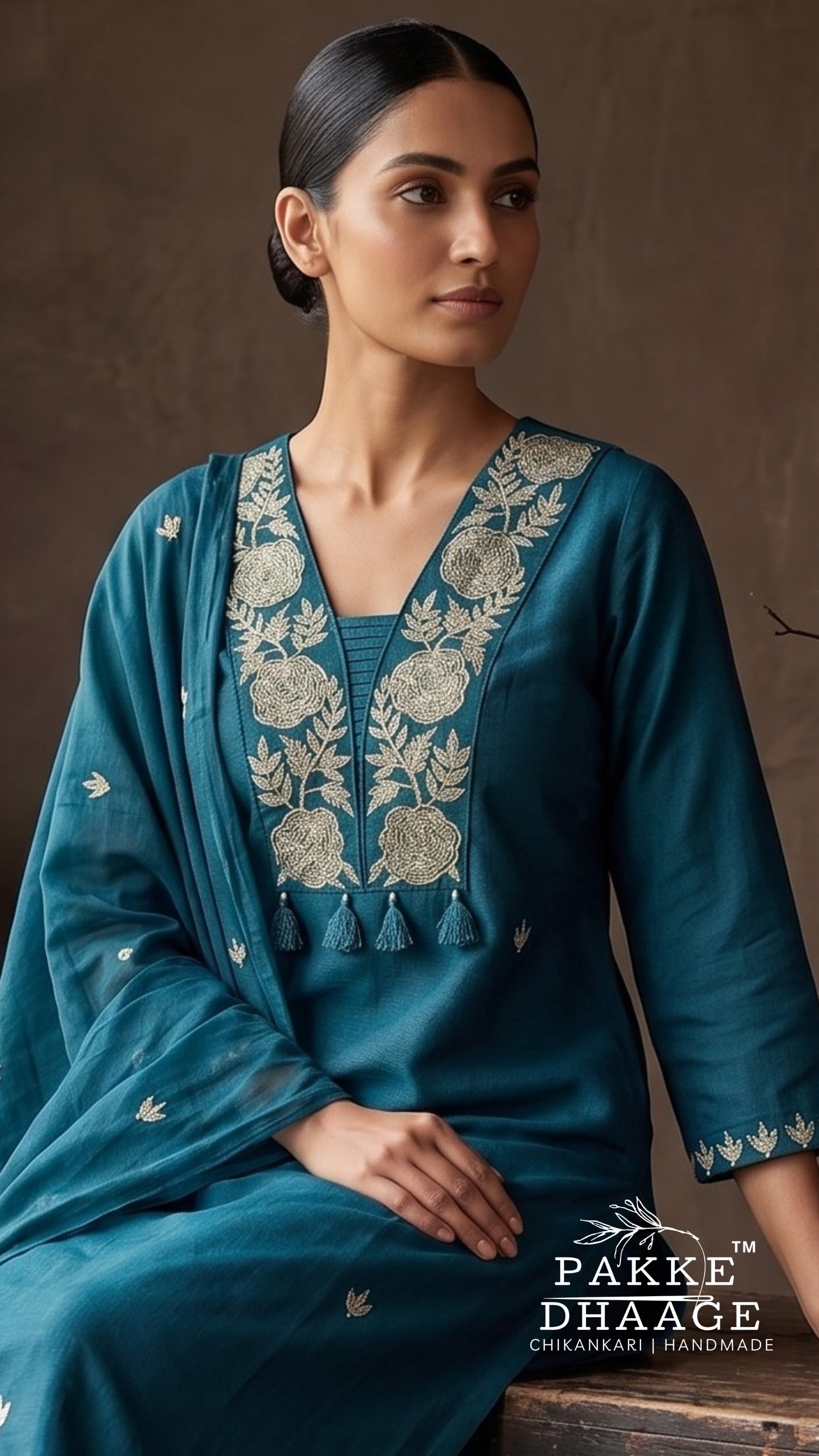 A woman wearing a teal blue embroidered kurta with a matching dupatta, featuring intricate cream floral embroidery on the neckline and sleeve cuffs. The brand logo 'PAKE DHAAGE' is visible in the bottom right corner.