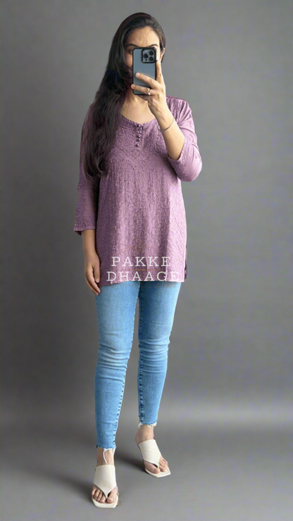Person taking a mirror selfie wearing a chikankari purple top and blue jeans in a room with plants.