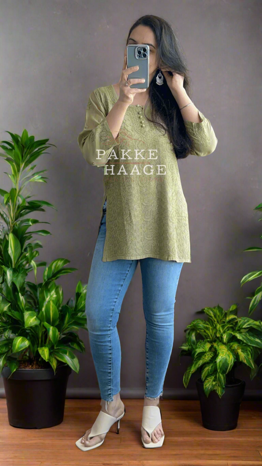 Person taking a mirror selfie wearing a chikankari mehendi green top and blue jeans, with plants around.