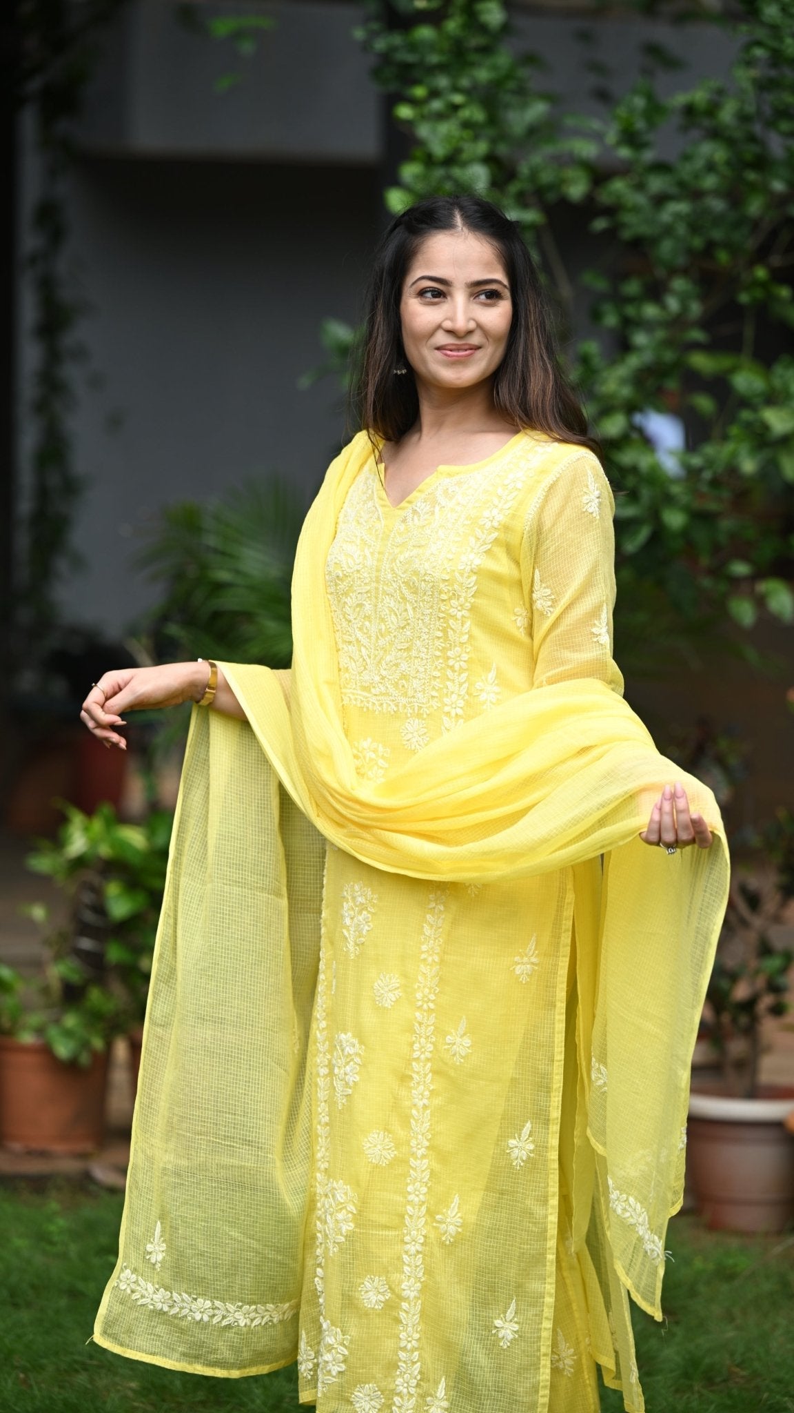 Woman in a yellow traditional outfit standing outdoors with greenery in the background