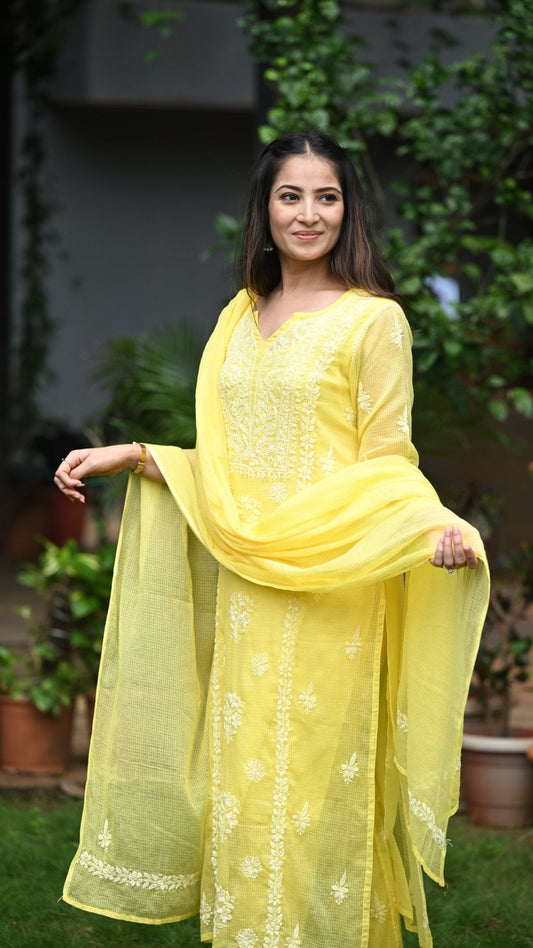 Woman in a yellow traditional outfit standing outdoors with greenery in the background