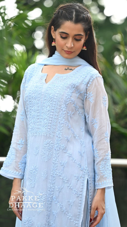 A woman wearing a light blue chikankari embroidered kurta with a matching dupatta, photographed outdoors against a blurred green foliage background. The brand name 'PAKE DHAAGE' is visible as a watermark in the bottom left corner.