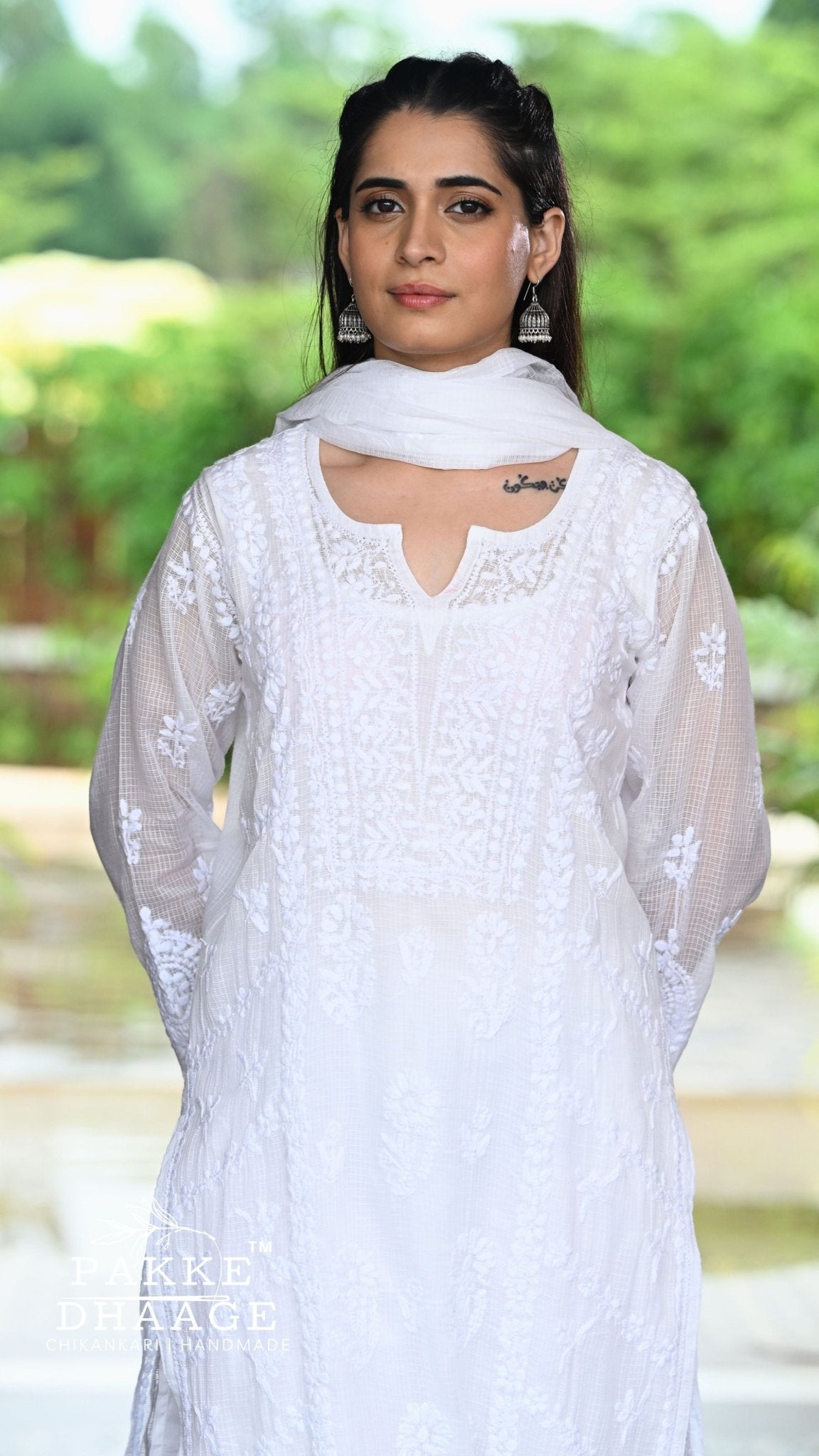 A woman wearing a white chikankari outfit with a matching white dupatta, photographed outdoors against a blurred green garden background. A 'Pakke Dressage' watermark is visible in the lower-left corner.
