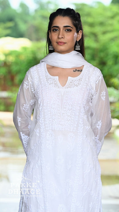 A woman wearing a white chikankari outfit with a matching white dupatta, photographed outdoors against a blurred green garden background. A 'Pakke Dressage' watermark is visible in the lower-left corner.