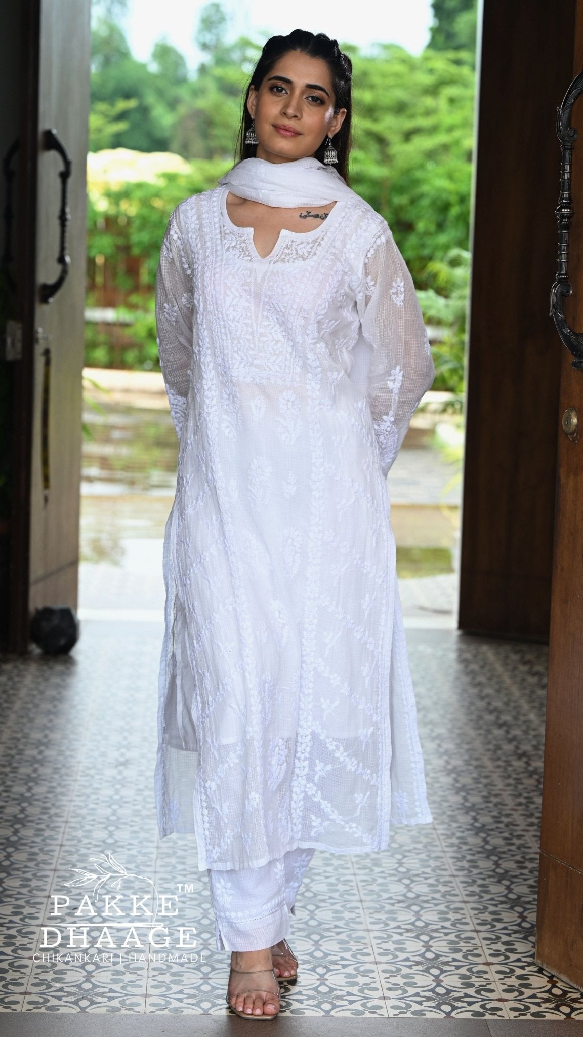 A woman wearing a white chikankari embroidered traditional outfit with a matching dupatta, standing in a doorway with a tiled floor and green garden background. The brand name 'Pakke Dhaage' is visible as a watermark in the bottom left corner.