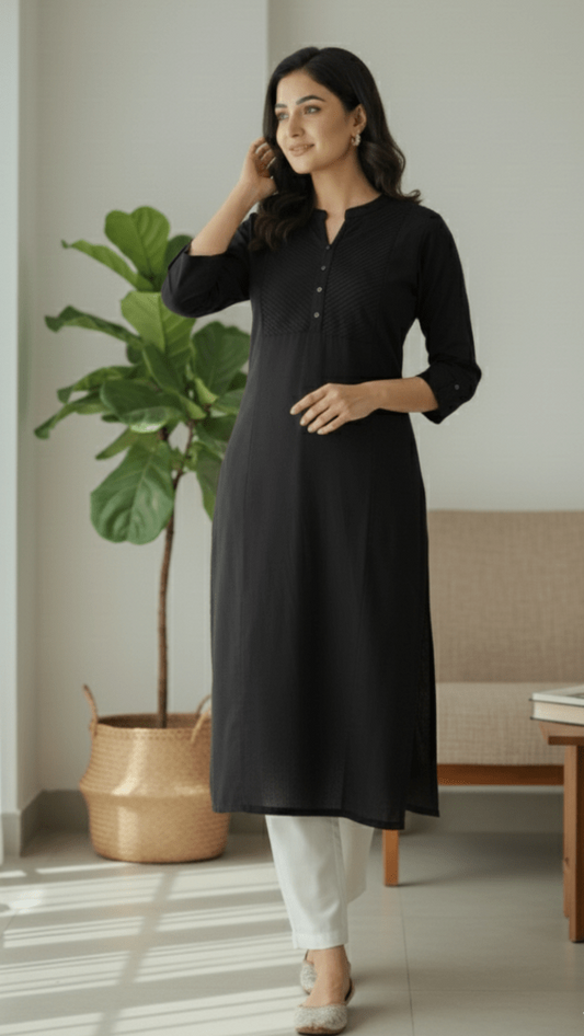 Woman wearing a black kurta with white pants in a room with a plant and furniture.