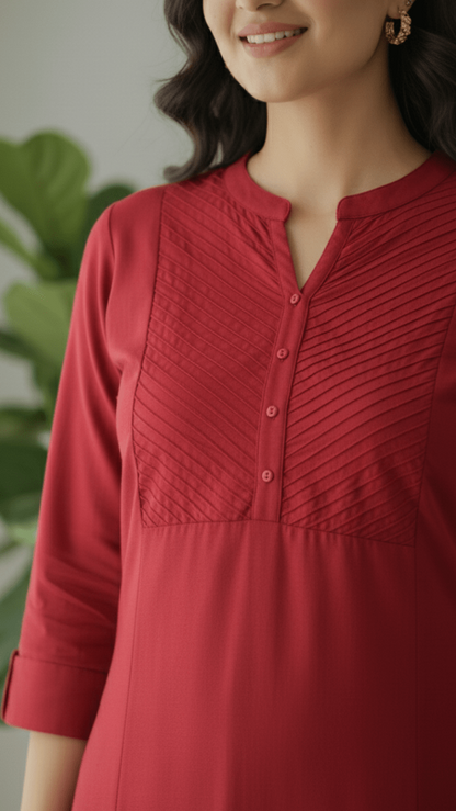 Woman wearing a red kurti with a pleated / pintex design, standing against a blurred green background.