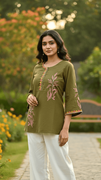 Woman in a green embroidered top and white pants standing in a garden.