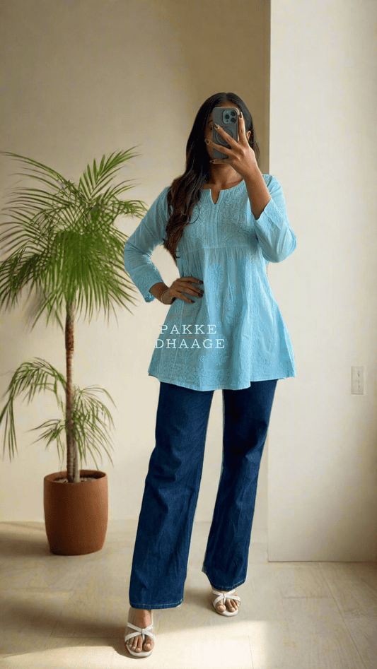 A woman standing on a balcony wearing a turquoise blue short kurti with Chikankari embroidery, paired with blue jeans and pink sandals.