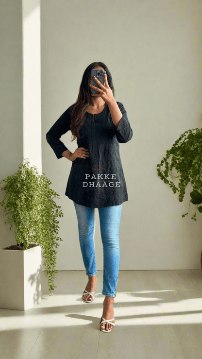 A person wearing a classic black short kurti with Chikankari embroidery, paired with blue jeans, standing in a room with a lot of plants.