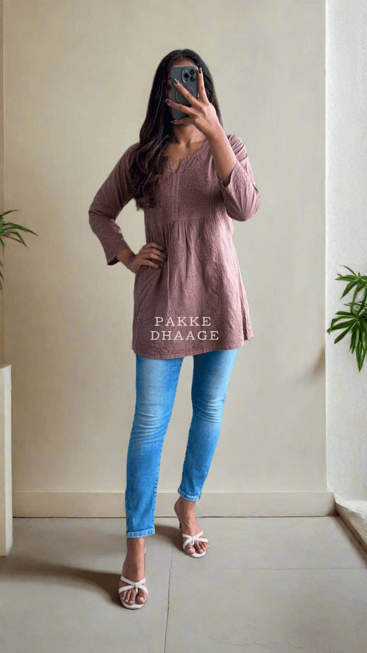 A person wearing a cocoa brown short kurti with Chikankari embroidery, paired with blue jeans and pink sandals.