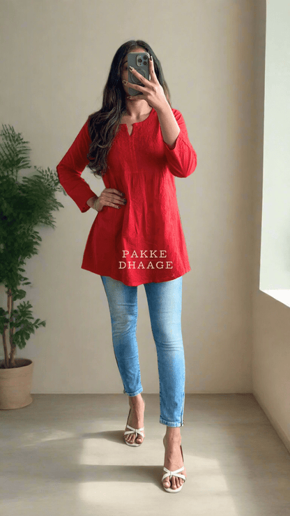 Person wearing a red top and blue jeans taking a mirror selfie in a plant-filled room.