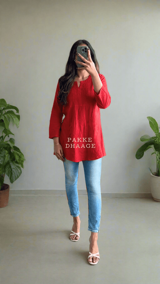 Person in a red shirt and blue jeans standing in a room with plants.