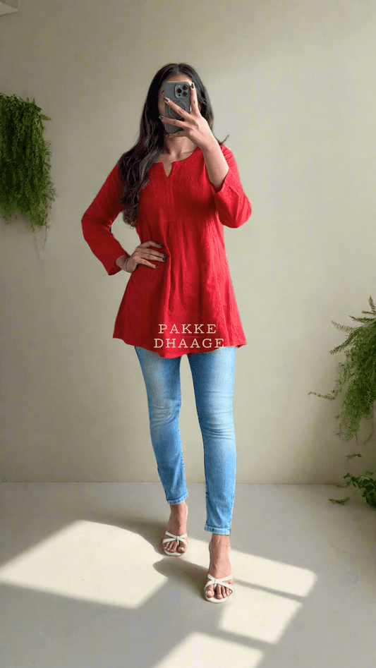 A woman standing in a room with plants, wearing a deep red short kurti with Chikankari embroidery, paired with blue jeans and beige sandals.
