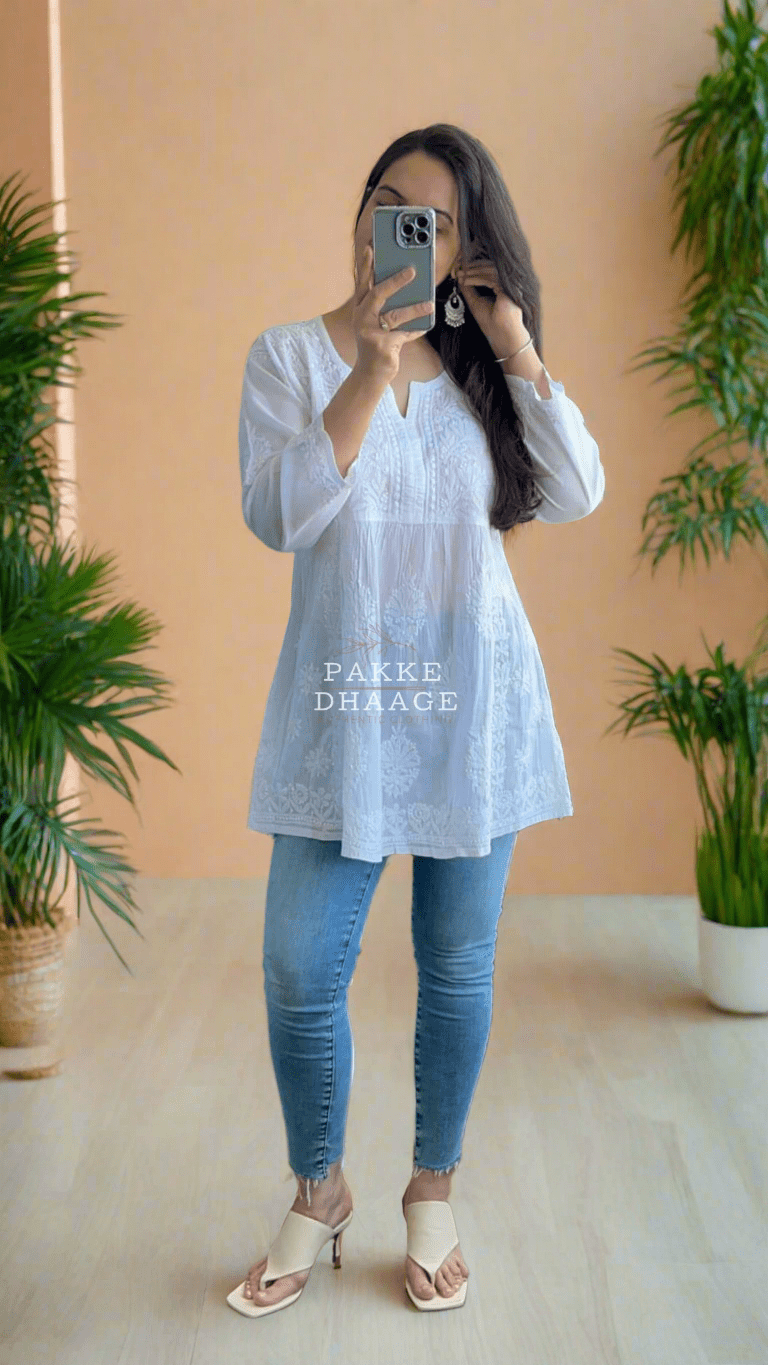 A woman standing on a balcony wearing a white short kurti with Chikankari embroidery, blue jeans, and heels.