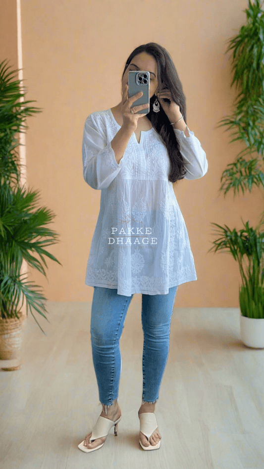 A woman standing on a balcony wearing a white short kurti with Chikankari embroidery, blue jeans, and heels.