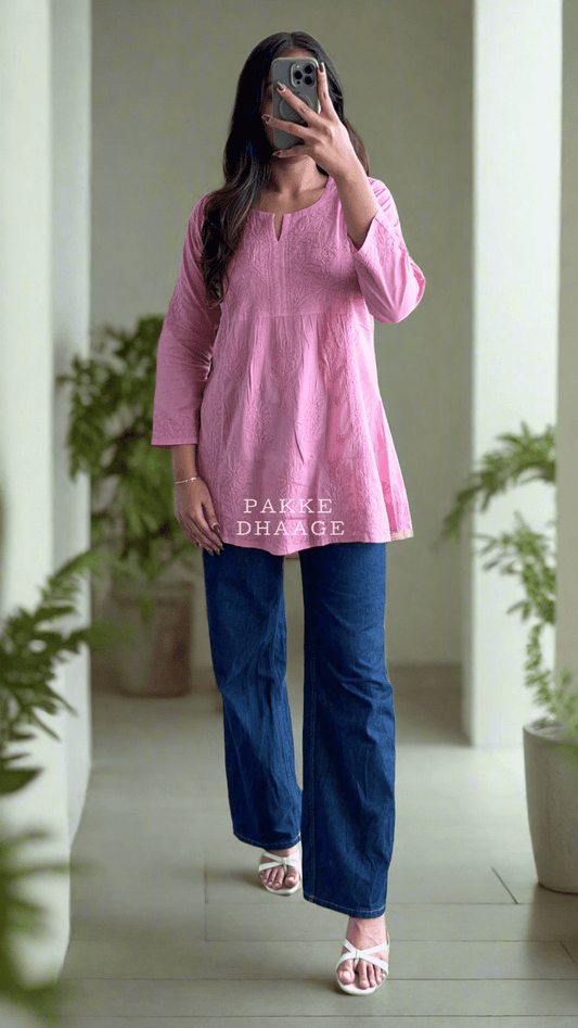 A woman standing on a balcony wearing a pink short kurti with Chikankari embroidery, paired with blue jeans and white sandals.
