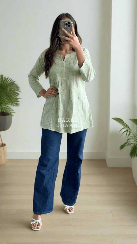 A woman wearing a pista green short kurti with Chikankari embroidery, paired with blue jeans and pink sandals.