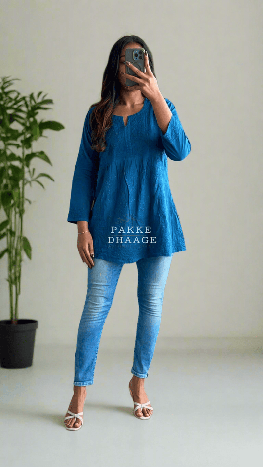 A person wearing a short blue kurti with Chikankari embroidery, paired with blue jeans and pink sandals, taking a mirror selfie.