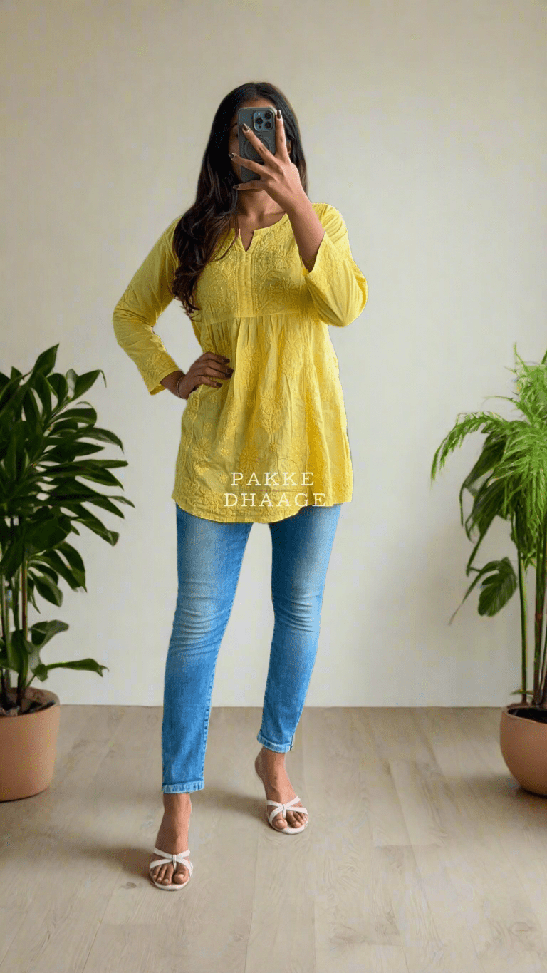 Person wearing a yellow top and blue jeans taking a mirror selfie in a room with plants.